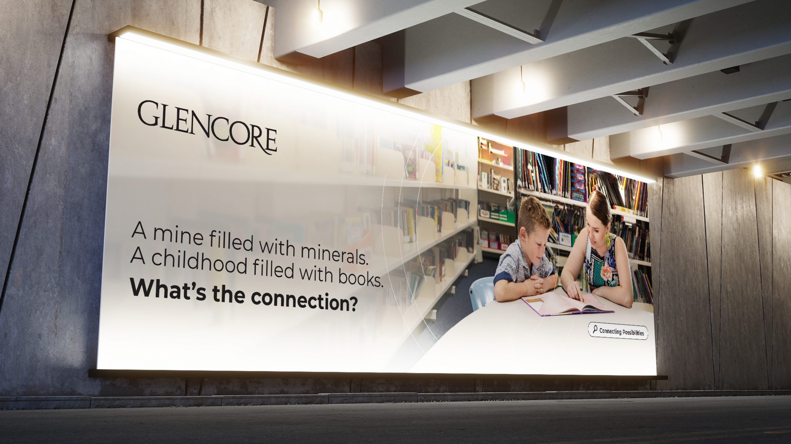 A billboard for the glencore advertising campaign with a woman and child sitting together in a library reading a book. The copy reads "a mine filled with minerals. A childhood filled with books. Whats the connection?"